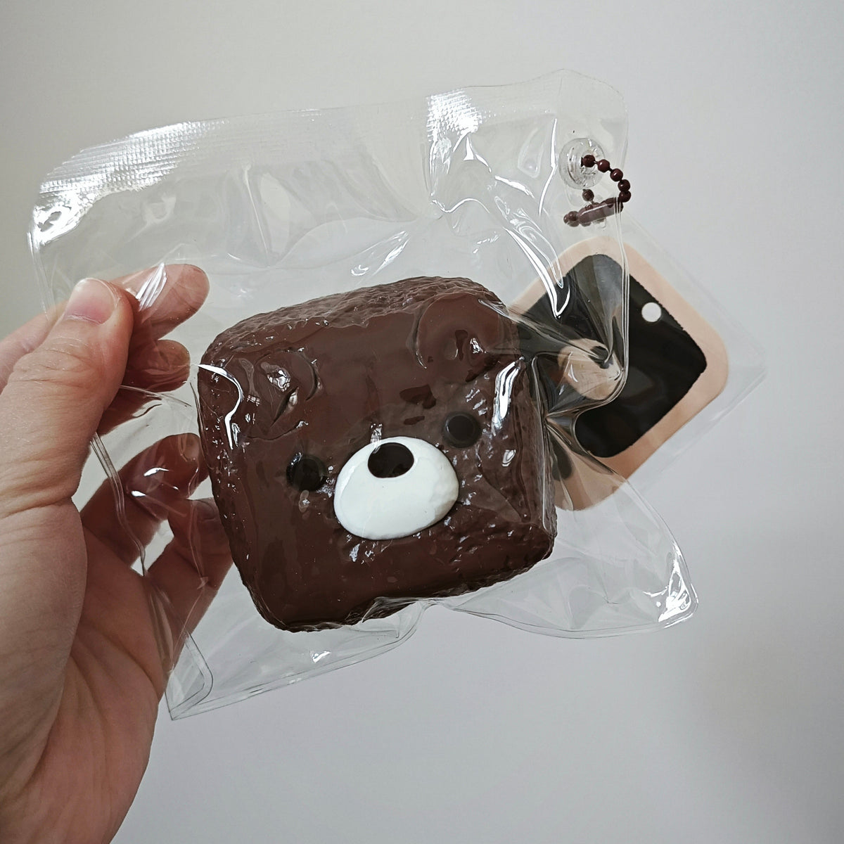 A hand holding a clear plastic bag containing a brown bear-face phone case with a white snail charm attached, against a plain light gray background.