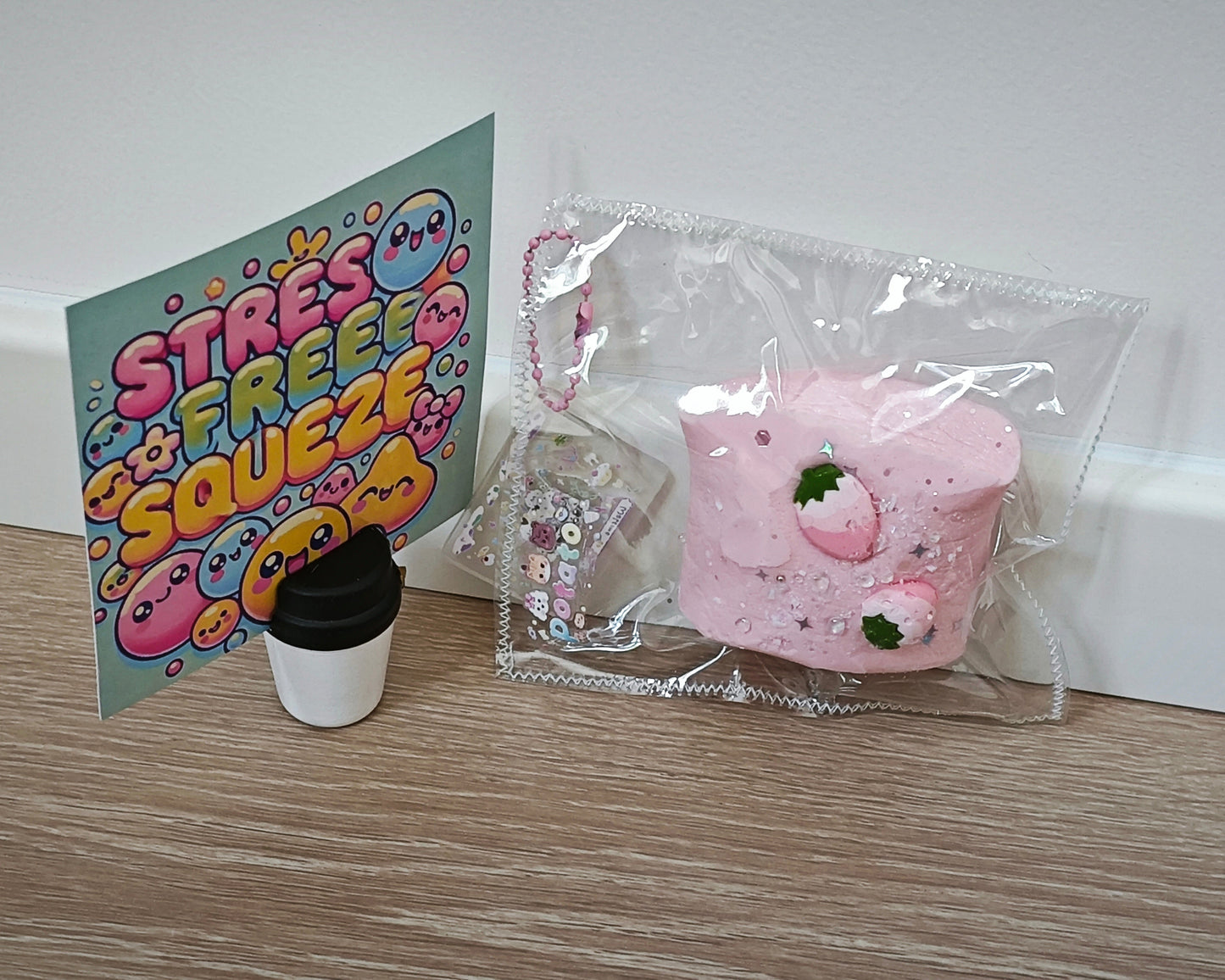 A pink strawberry-themed squishy toy in a clear plastic bag sits on a light wood surface next to a colorful 'STRESS FREE SQUEEZE' card and a small black-and-white cup-shaped accessory.