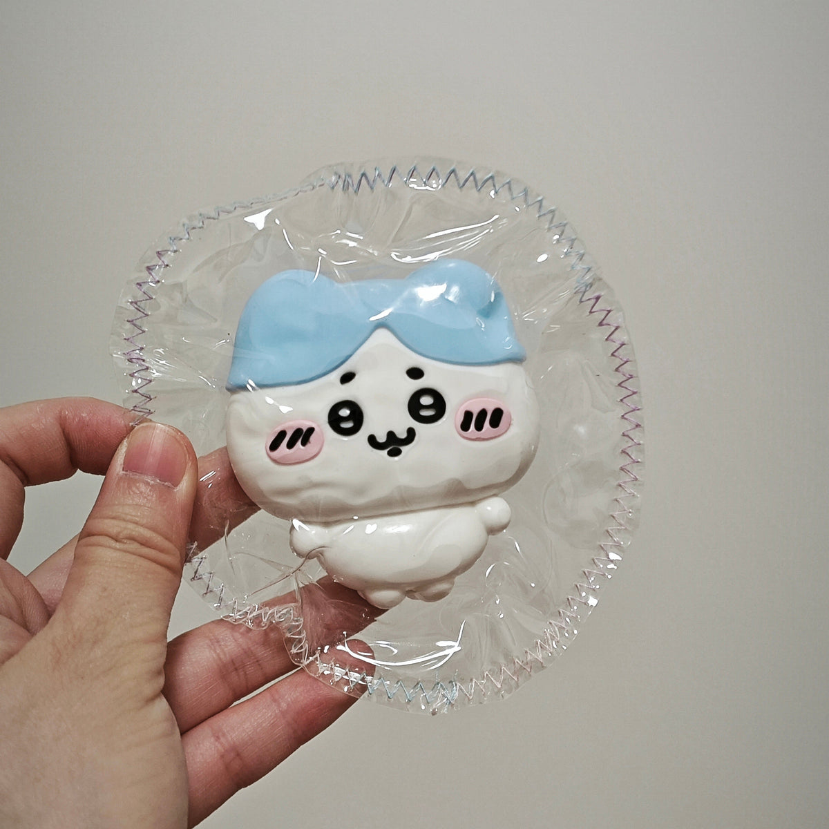 A hand holding a small white cartoon figurine with a light blue bow and black facial features, still in clear plastic wrapping, against a plain light gray background.