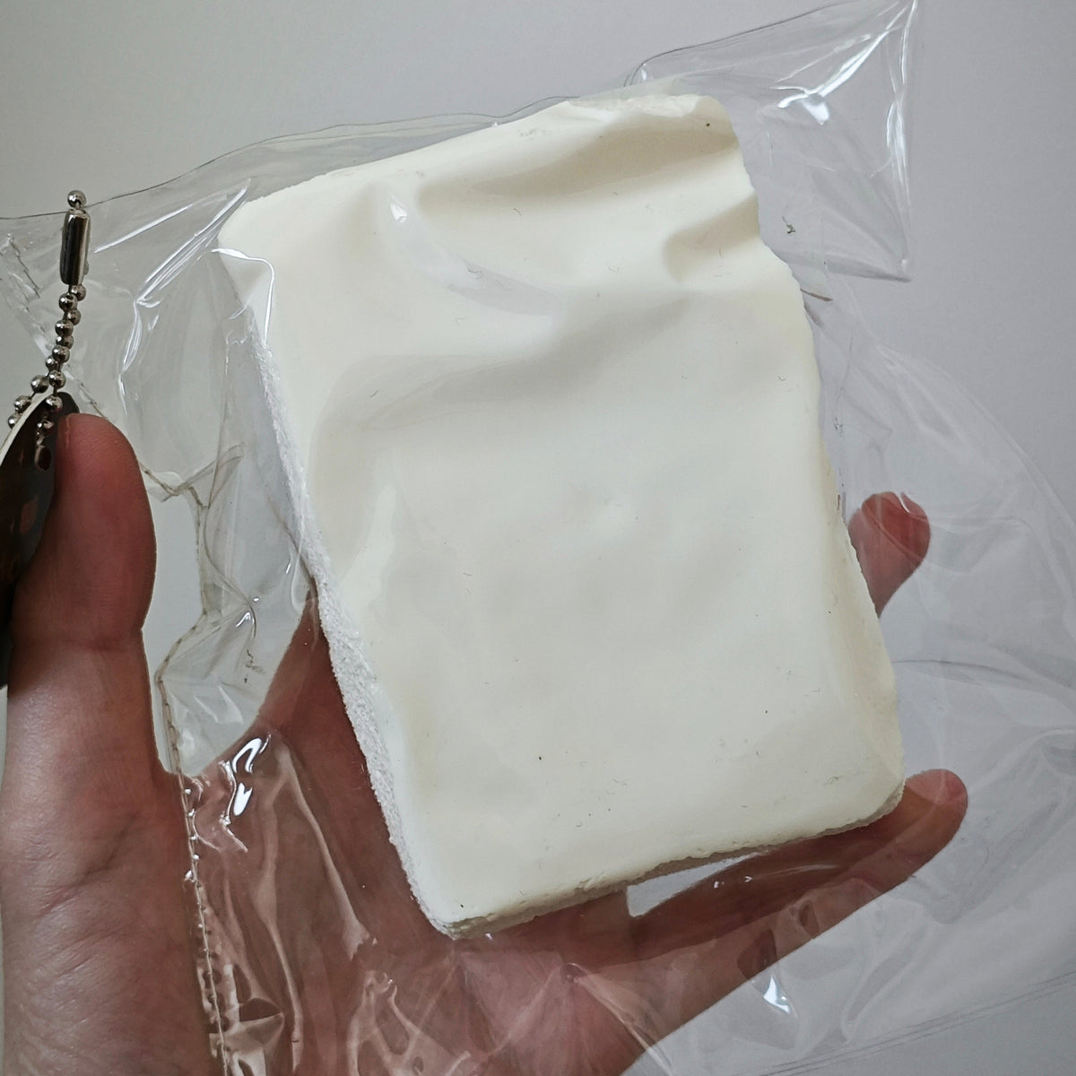 A hand holding a white rectangular foam block or sponge wrapped in clear plastic packaging against a plain light gray background.