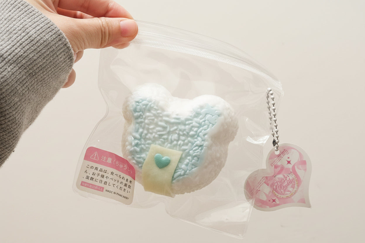 A hand holds a clear zip bag containing a soft, fuzzy bear-shaped item in white and pale blue with a small light blue heart detail, alongside a pink heart-shaped charm on a silver chain, against a plain light background.