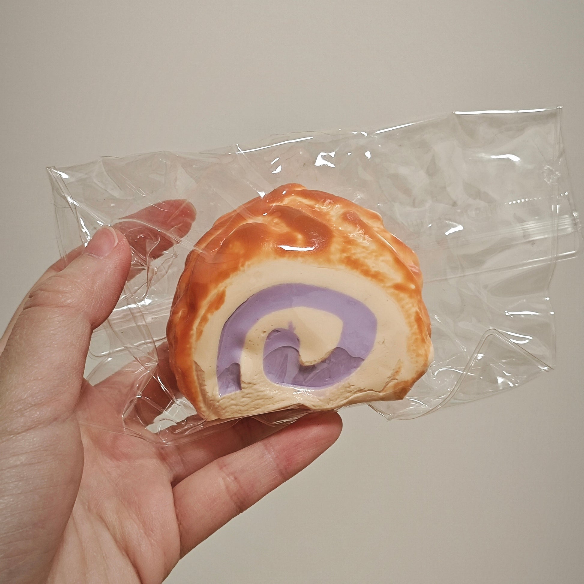 A hand holding a small, round, pastry-like item wrapped in clear plastic. The item has a golden-brown outer surface and a spiral cross-section showing cream and purple layers, set against a plain light beige background.