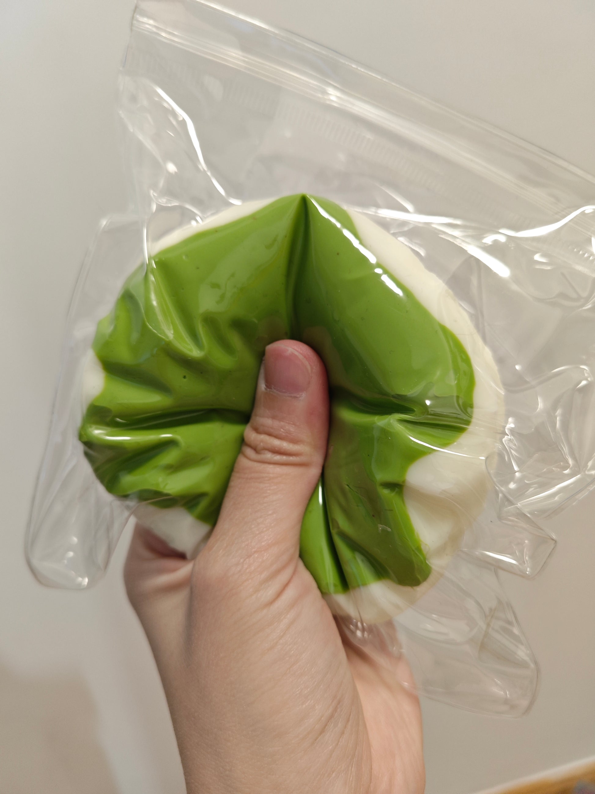 A hand holding a green and white ruffled silicone or rubber item inside a clear plastic bag against a plain light background.