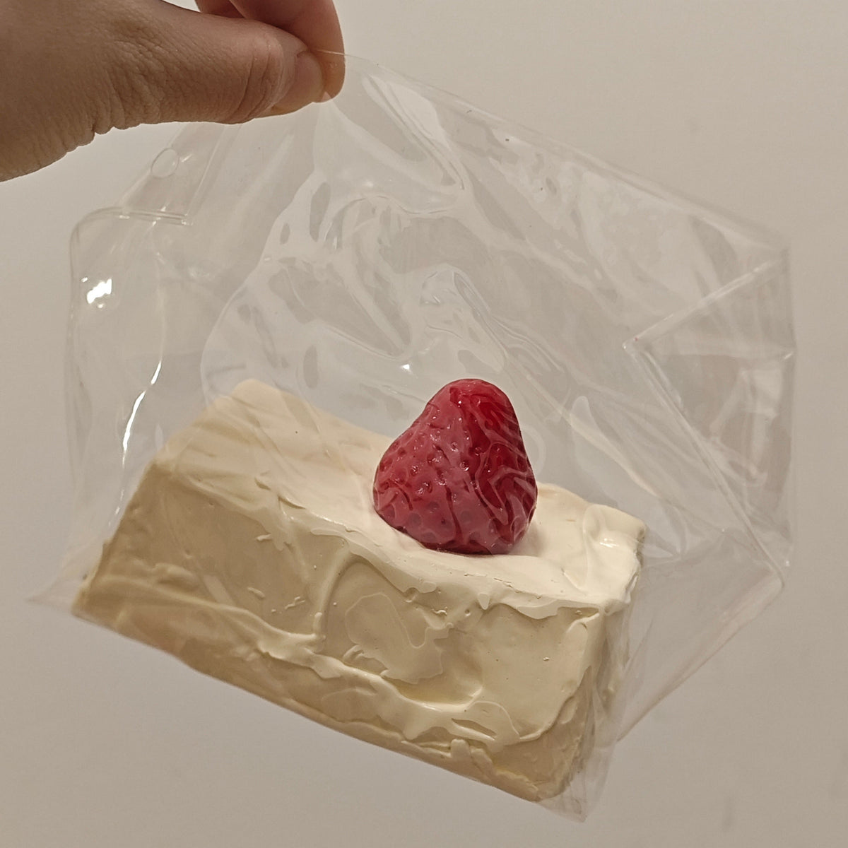 A hand holding a clear plastic bag containing a small white rectangular item topped with a red strawberry, against a plain light background.