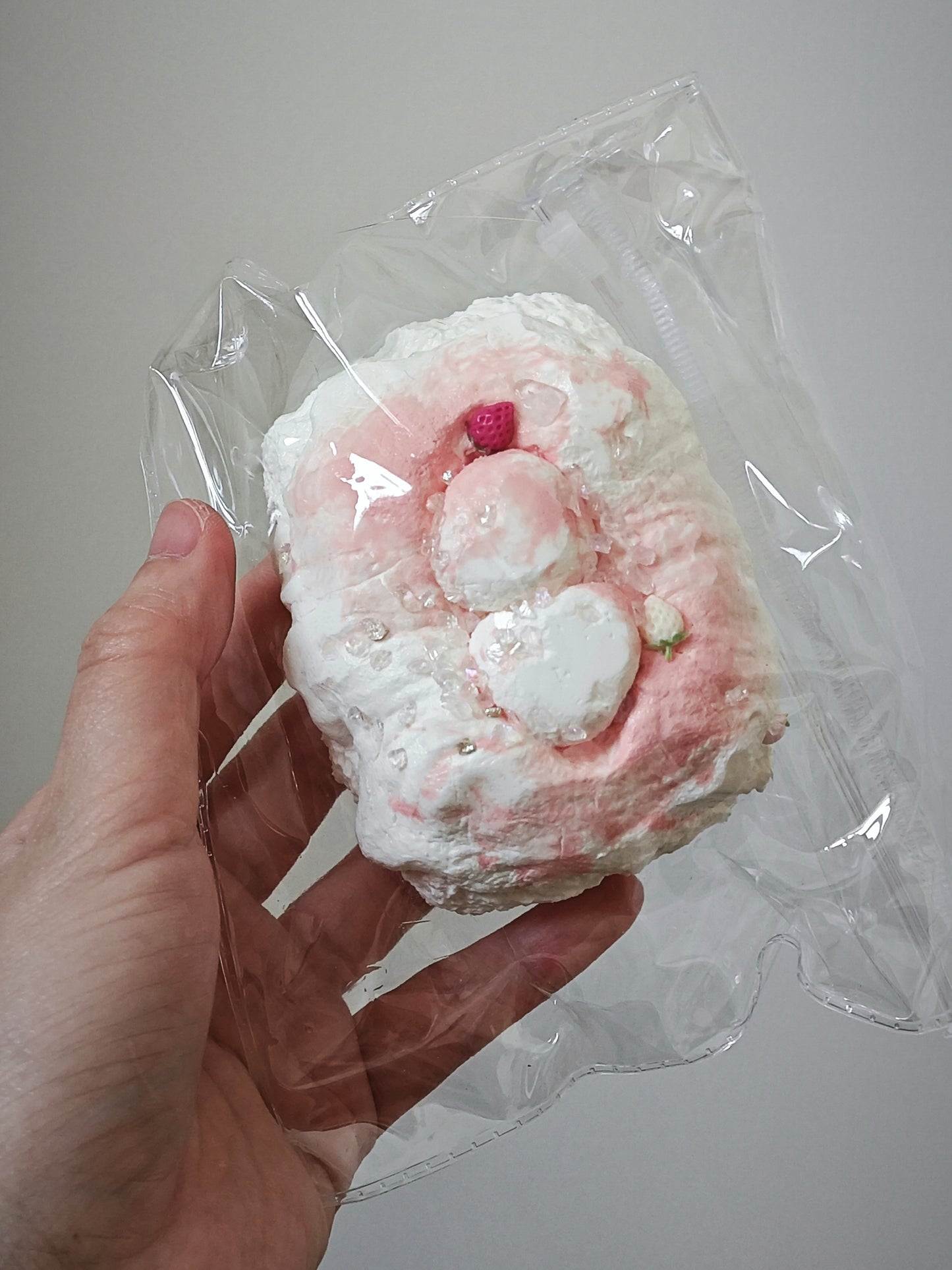 A hand holding a clear plastic bag containing a pink and white textured, marshmallow-like novelty item against a plain light gray background.
