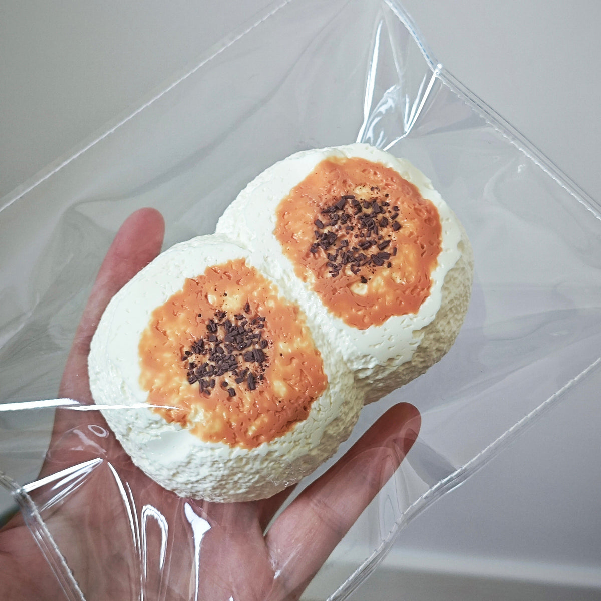 Two round white pastries with orange-brown centers and dark sprinkles, held in a hand inside a clear plastic bag against a plain indoor background.