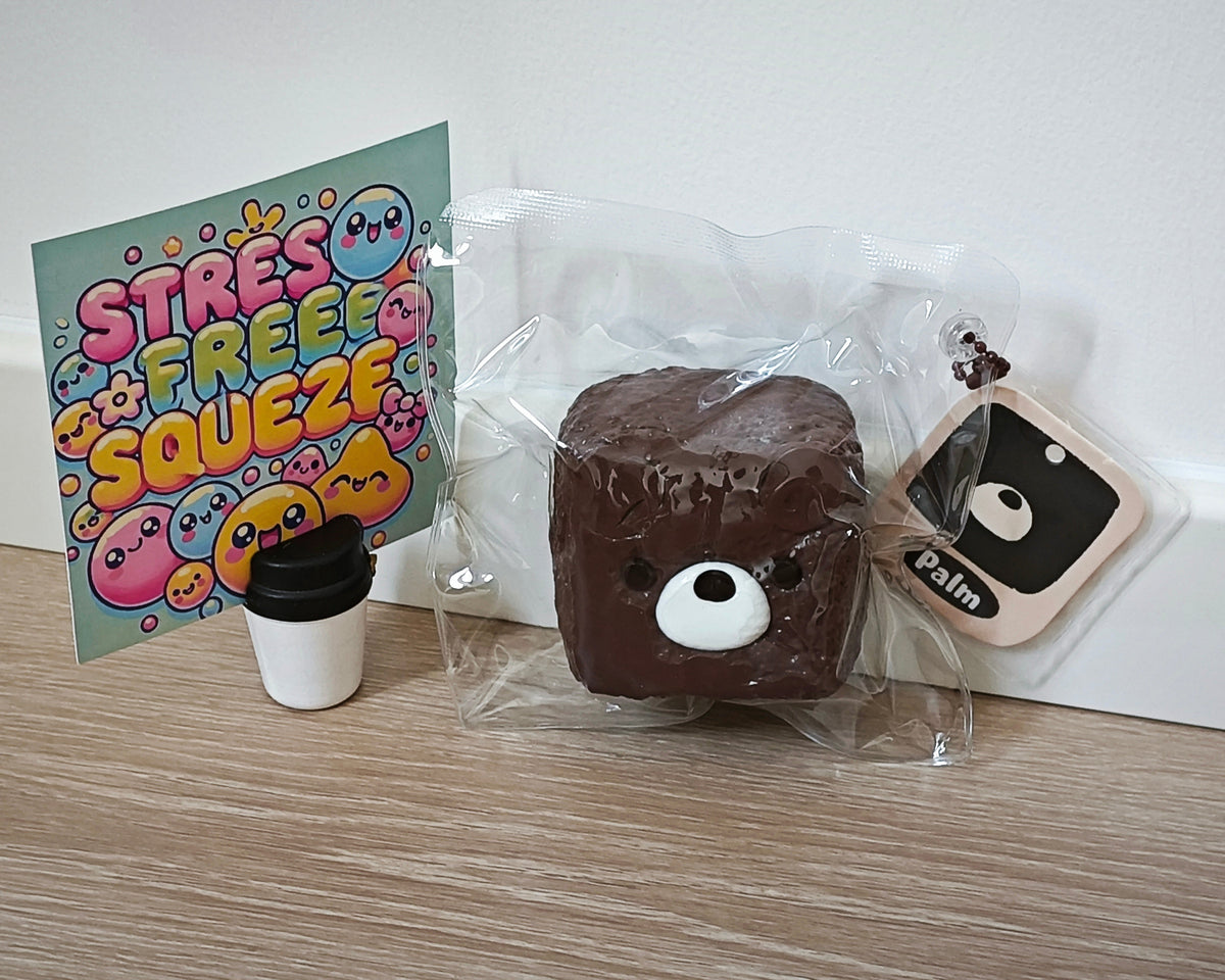 A square brown squishy toy with a bear face, sealed in clear plastic packaging, displayed on a light wood surface against a white wall. A colorful 'STRESS FREE SQUEEZE' card, a small coffee cup charm, and a phone-shaped tag labeled 'Palm' are placed beside it.