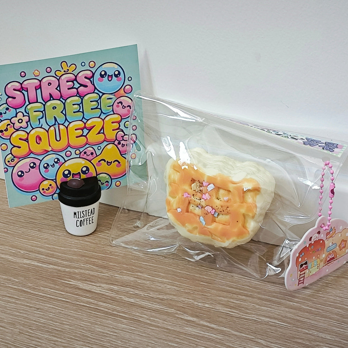 A squishy toy shaped like a pastry or bread, sealed in a clear plastic bag, displayed on a wooden surface next to a small cup-shaped item and a colorful card that reads 'STRESS FREE SQUEEZE'.