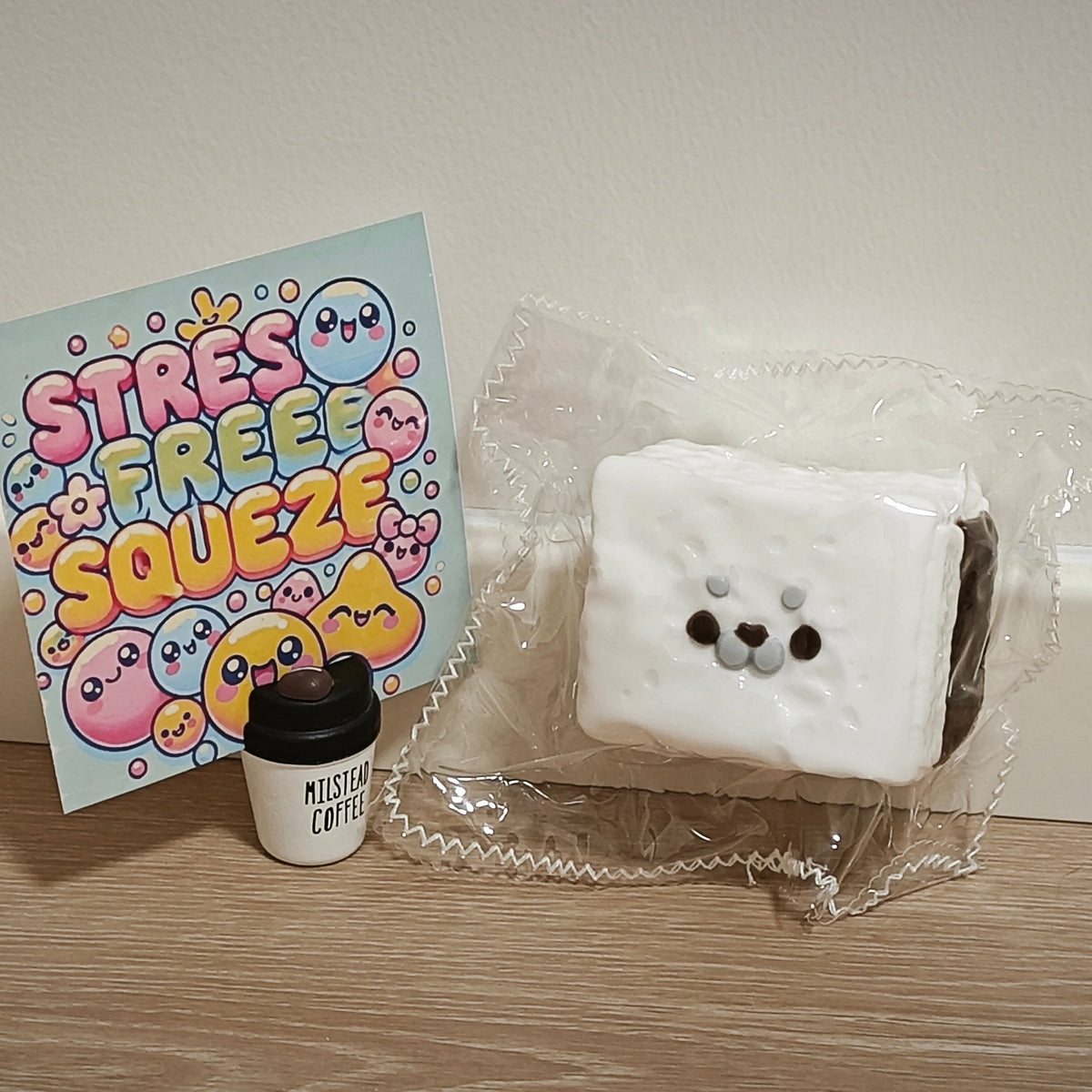 A white square squishy toy with a cute seal face, packaged in clear plastic, displayed next to a colorful 'Stress Free Squeeze' card and a small black coffee cup accessory on a wooden surface against a plain wall.