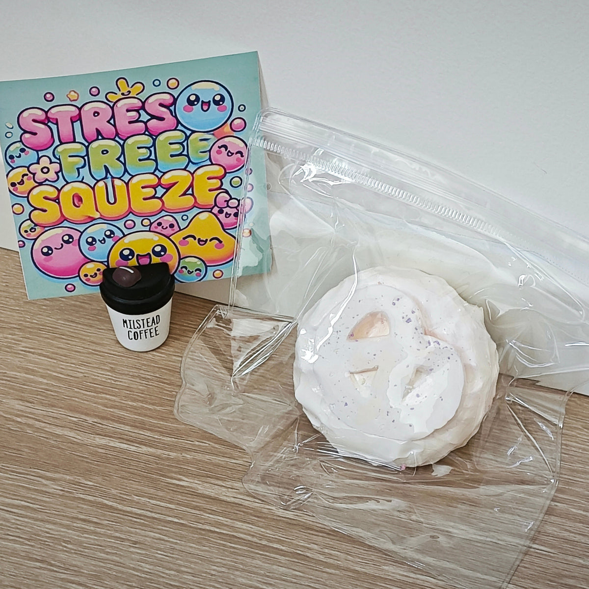 A white foam squishy toy sealed in a clear plastic bag, shown on a wood floor next to a small cup labeled 'PUDDLESTAR COFFEE' and a colorful card that reads 'STRESS FREE SQUEEZE'.