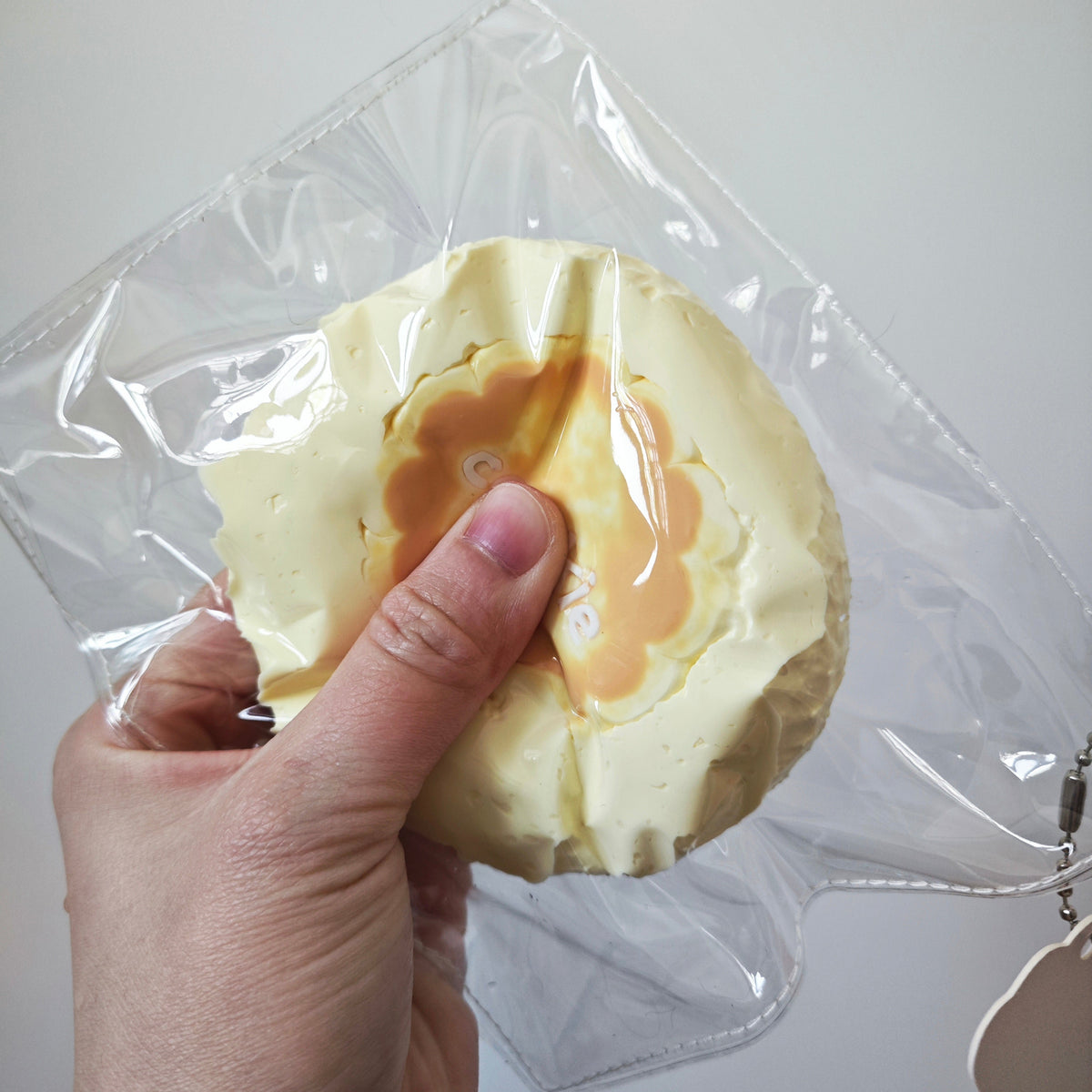 A hand holding a clear plastic zippered pouch with a round, pale yellow and orange textured insert or pouch inside, against a plain light gray background.