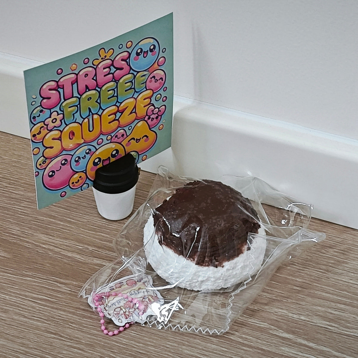 A novelty squishy toy shaped like a chocolate-covered ice cream sandwich in clear plastic packaging, displayed on a light wood floor next to a small coffee cup and a colorful 'Stress Free Squeeze' card.