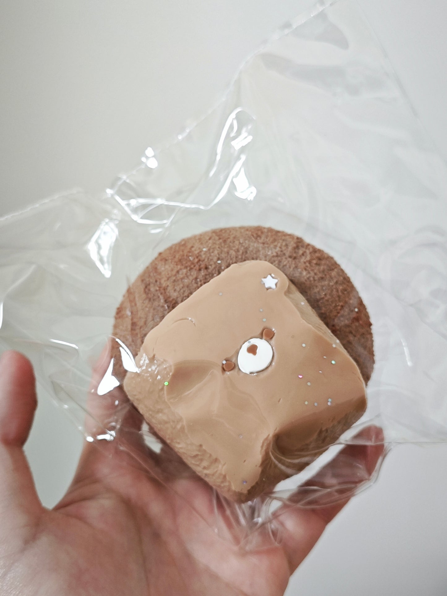 A hand holding a small round brown item wrapped in clear plastic, with a beige bear-face decoration on top.
