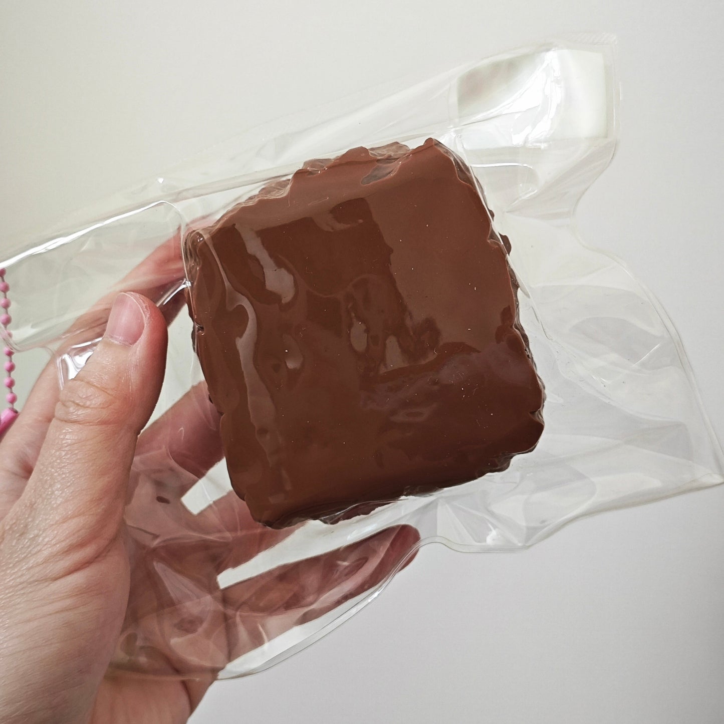 A hand holding a square brown chocolate-covered item inside a clear plastic bag against a plain light background.