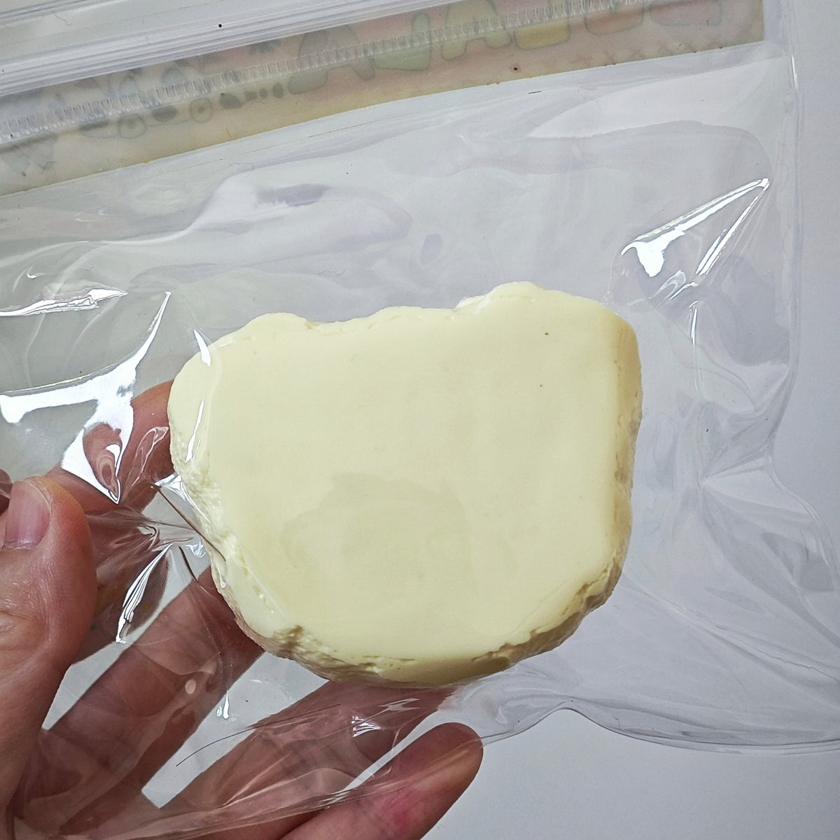 A hand holding a clear plastic zip-top bag containing a pale yellow, irregularly shaped foam or sponge-like object against a plain light background.