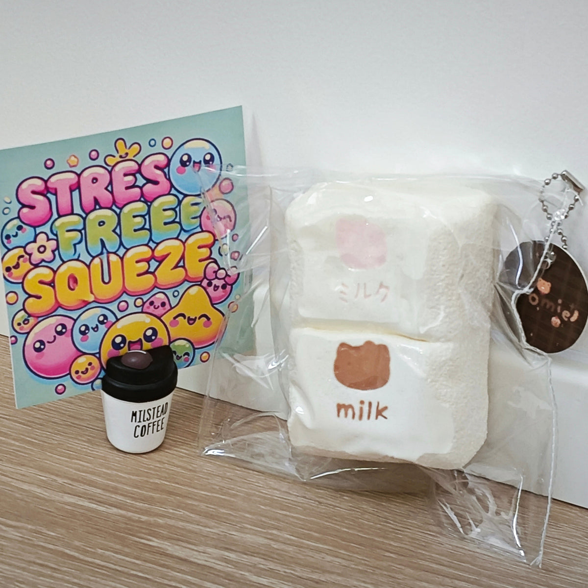 A set of cute squishy toys and accessories on a light wood floor against a white wall. The set includes a pastel 'STRESS FREE SQUEEZE' card, a small 'MISTLETOE COFFEE' cup, and a large clear bag containing two white marshmallow-shaped squishys labeled 'milk' with a brown bear face, plus a brown keychain.