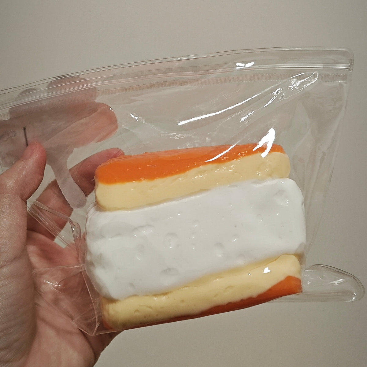 A hand holding a clear plastic zip bag containing a small sandwich-shaped item with orange and cream-colored layers, photographed against a plain light gray background.