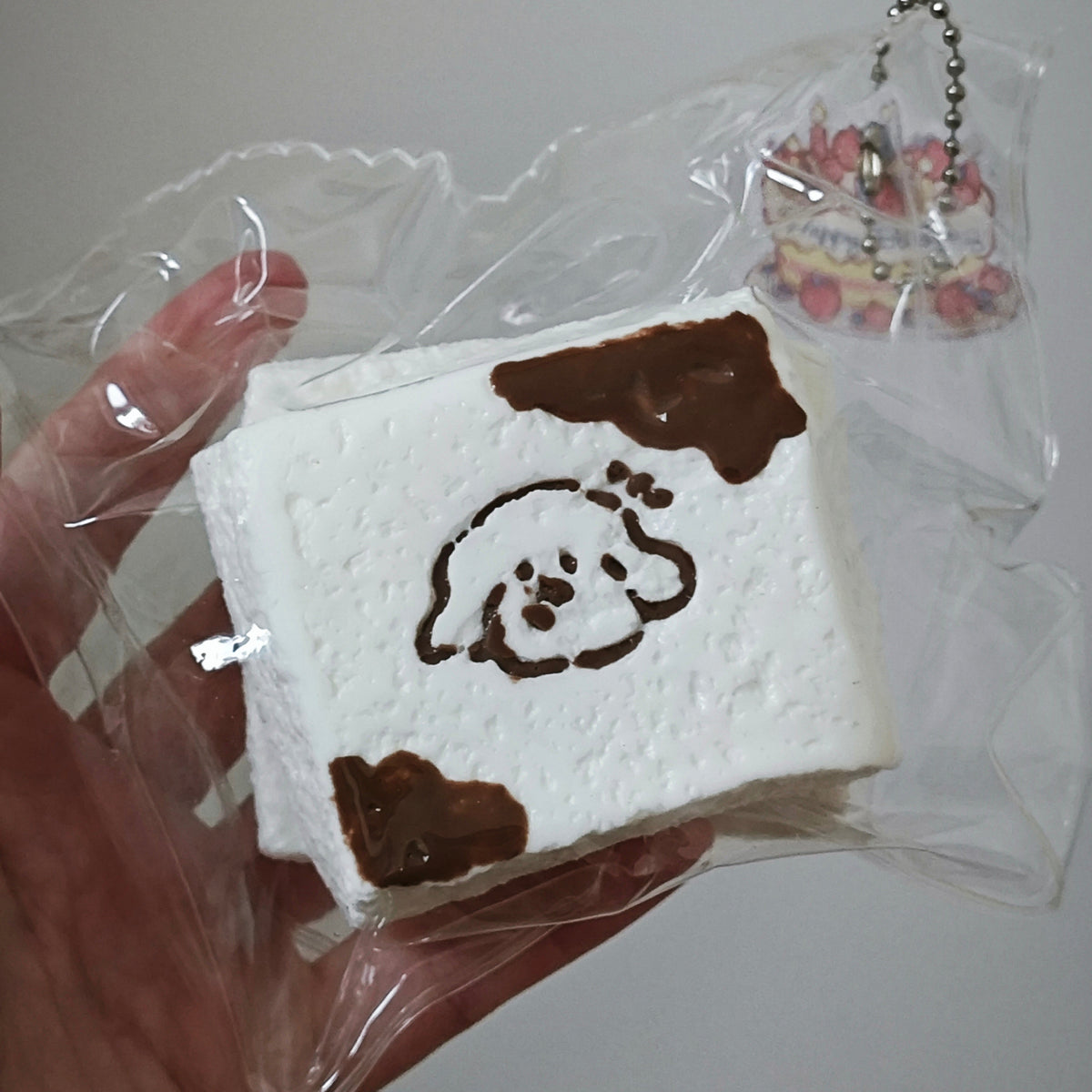 A hand holding a square white squishy toy with brown cow-print spots and a simple dog face illustration, sealed in clear plastic packaging with a small charm attached.