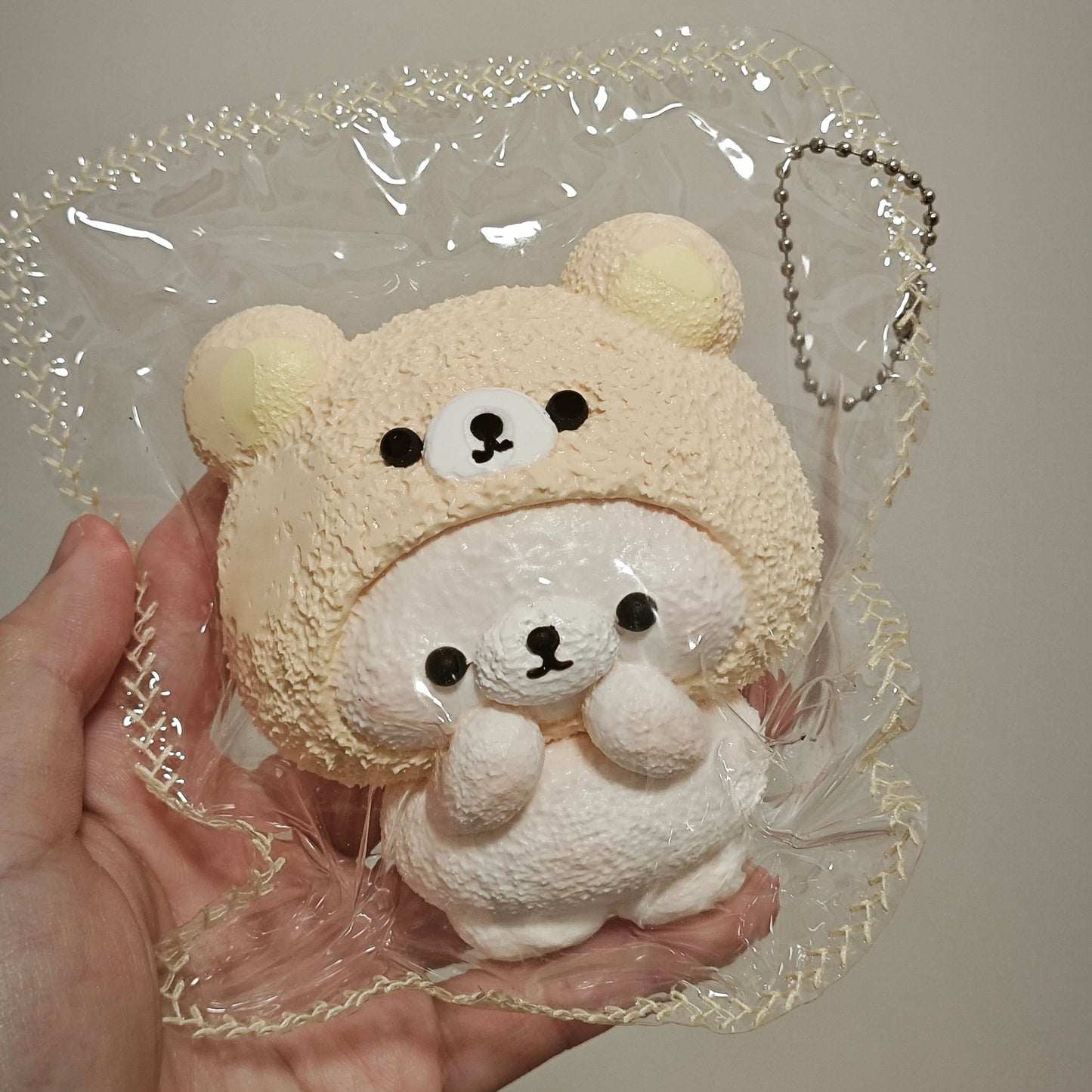 A hand holding a plush toy shaped like a bear wearing a beige hood, packaged in a clear plastic bag with a decorative trim and a metal keychain loop, against a plain light background.