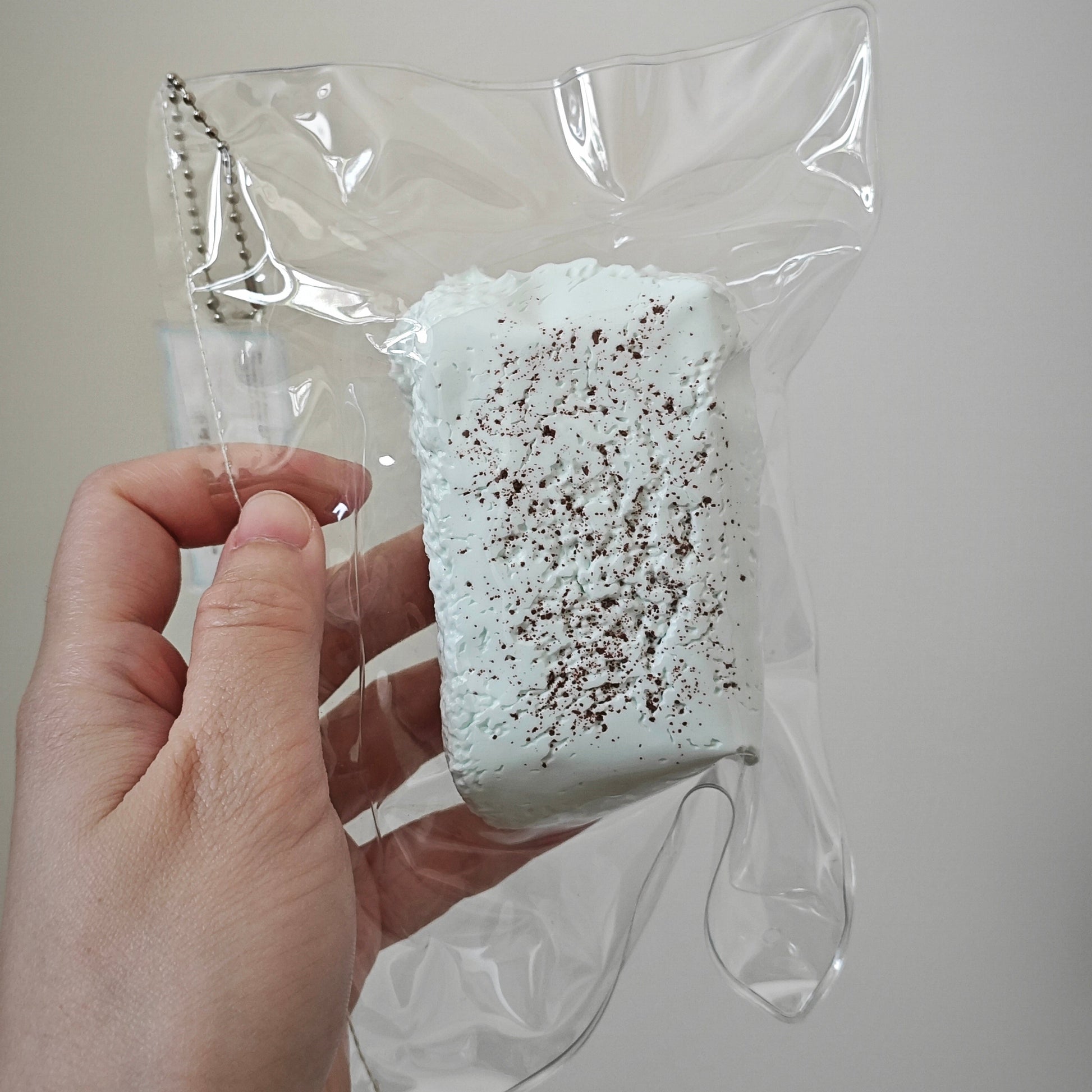 A hand holding a packaged squishy toy. The toy is a rectangular, light blue-green marshmallow-like item covered in dark brown sprinkles, sealed inside a clear plastic bag with a metal ball chain attached.