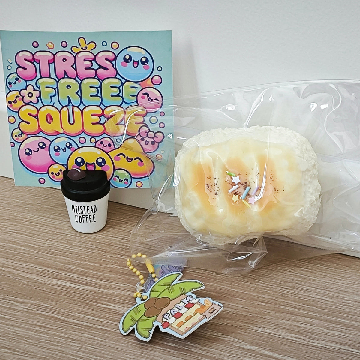 A stress-free squishy toy set on a light wood floor against a white wall, featuring a packaged cream-colored squishy, a small black coffee cup keychain with 'Mustead Coffee' text, and a colorful 'Stress Free Squeeze' card.