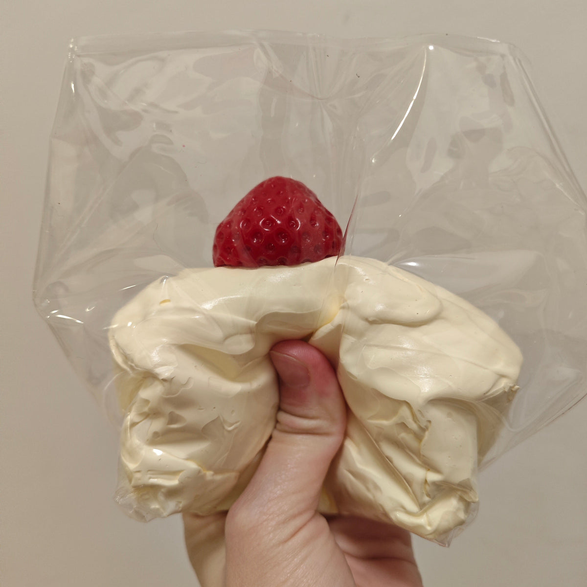 A hand holding a clear plastic bag containing a sculpted cream-colored object with a red berry on top, photographed against a plain light beige wall.