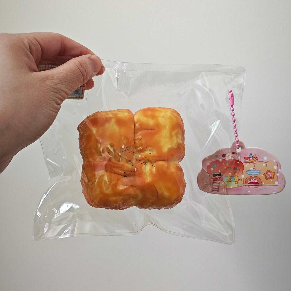 A hand holds a clear plastic bag containing a golden-brown, four-puck playdough or clay sculpture, with a small pink character charm attached on the right side, against a plain light gray background.