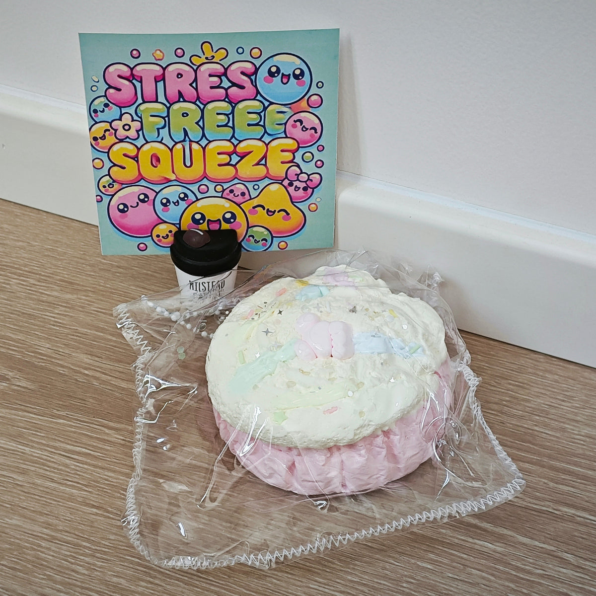 A pink and white frosted squishy toy wrapped in clear plastic sits on a light wood floor in front of a white wall, with a small black jar and a colorful 'STRESS FREE SQUEEZE' card behind it.