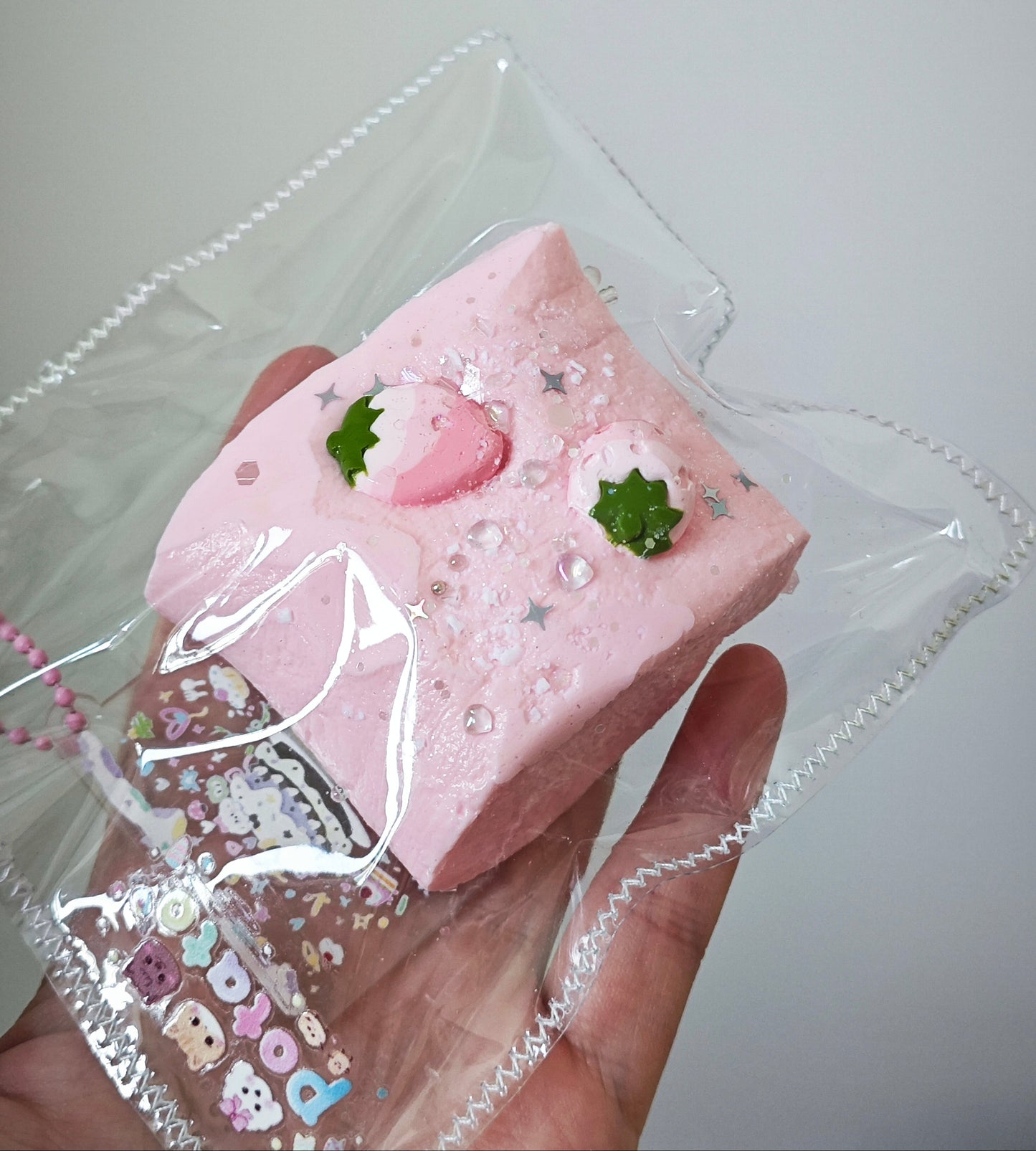 A hand holds a pink rectangular decorative item wrapped in a clear plastic bag. The item has a strawberry-themed design with green leaf accents, small silver star glitter, and a small Hello Kitty charm attached near the bottom.
