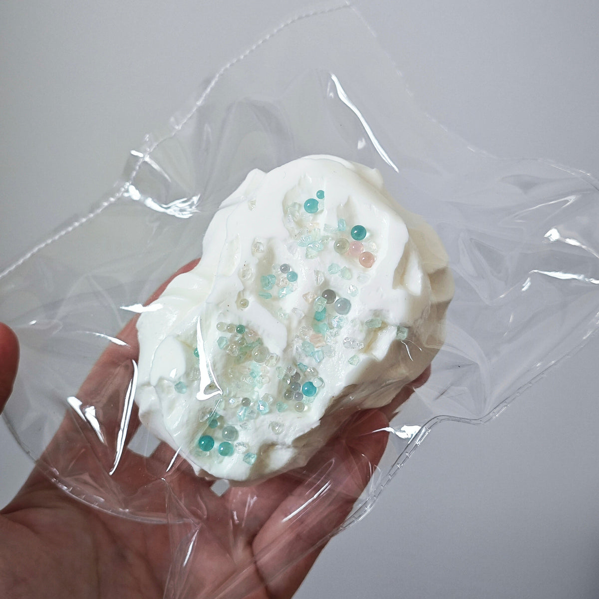 A hand holding a clear plastic bag containing a white, textured, gel-like squishy toy with small translucent blue and pink beads, against a plain light gray background.