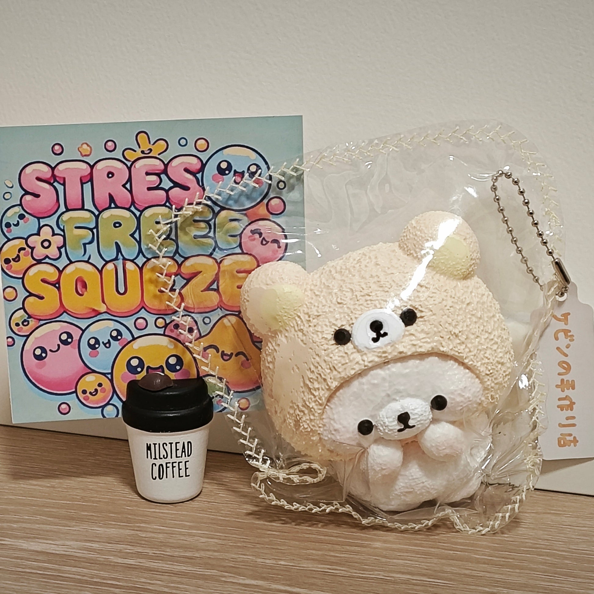 A cute plush bear keychain in a clear plastic bag, shown next to a small white coffee cup and a colorful 'Stress Free Squeezie' card on a wooden surface against a white wall.