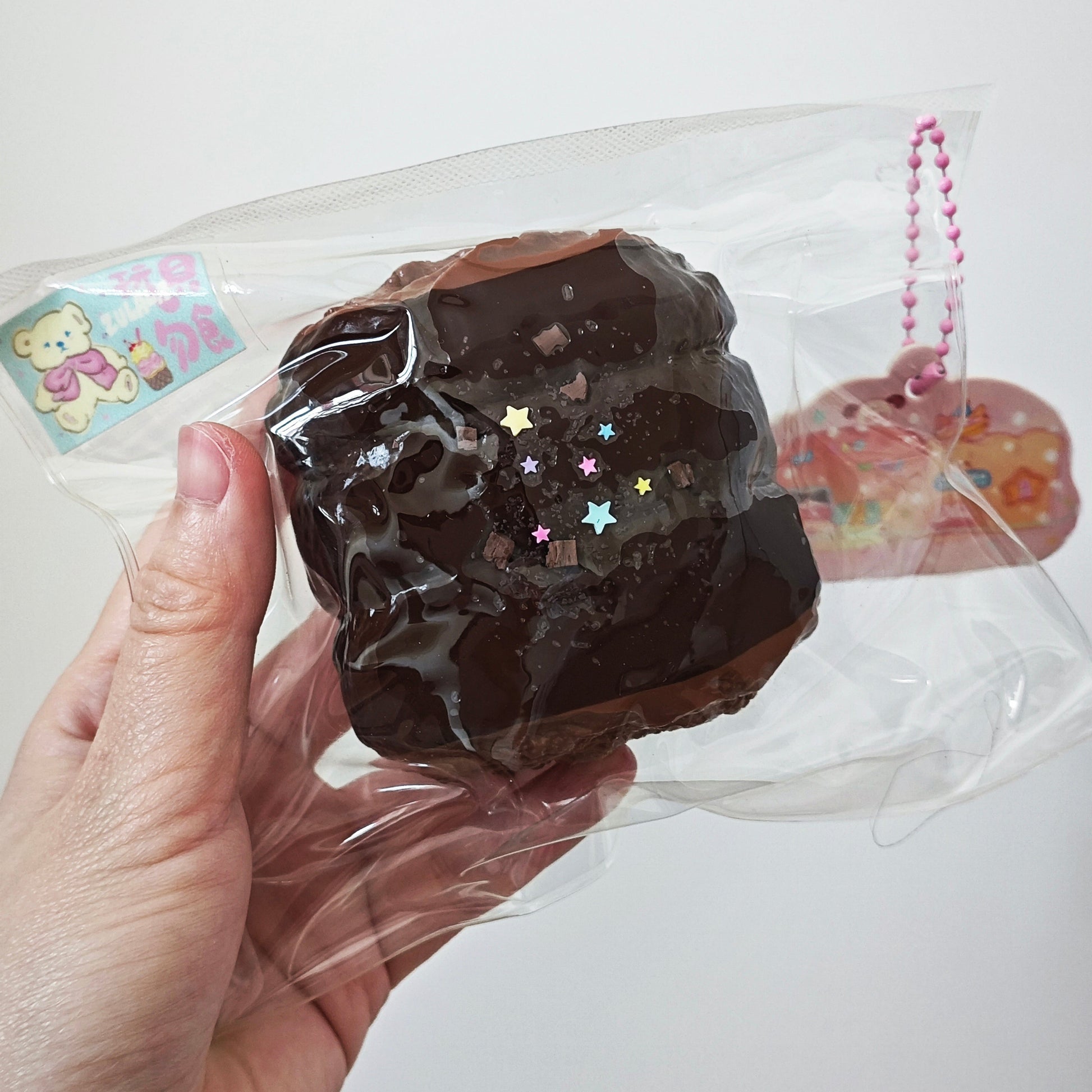 A hand holding a clear plastic bag containing a brown chocolate-themed squishy toy with small multicolored star accents. A pink beaded charm is attached on the right side, and a small bear sticker is visible on the left.