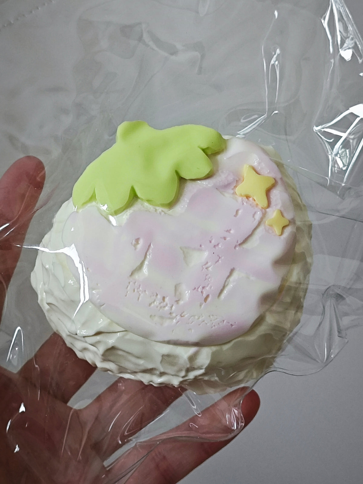 A hand holding a round, strawberry-themed squishy toy wrapped in clear plastic. The toy features a pale pink top with embossed details, a light green strawberry leaf, and two small yellow stars, set against a neutral gray background.