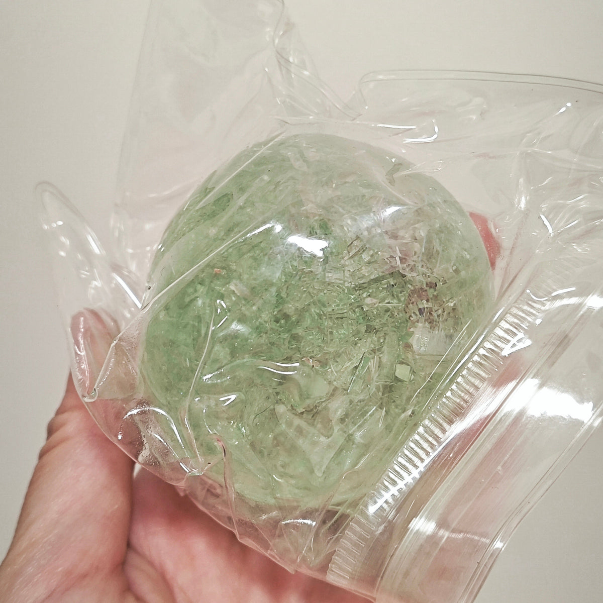 A hand holding a small, round, translucent light green item sealed inside a clear plastic bag against a plain light background.