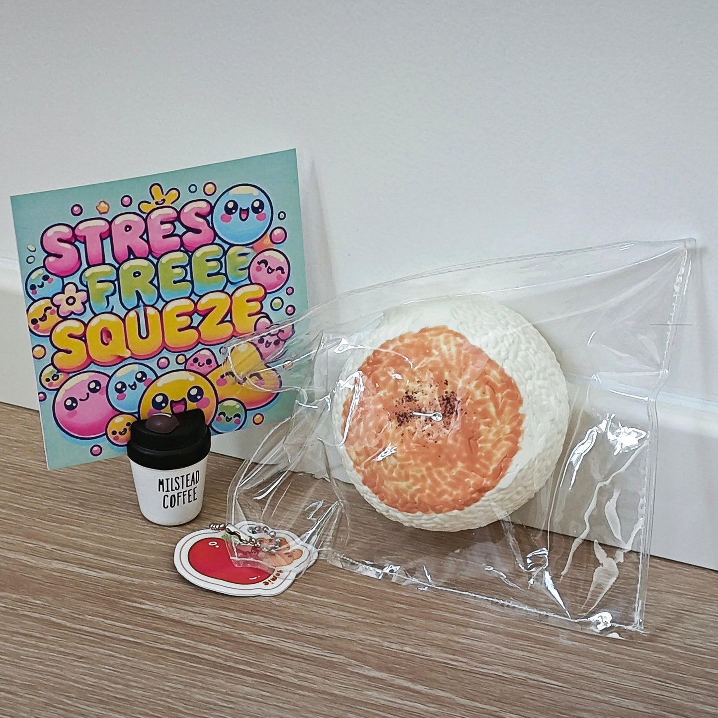 A stress relief gift set on a light wood floor against a white wall, featuring a 'Stress Free Squeeze' card, a miniature 'Mister Coffee' cup, a red heart-shaped sticker, and a peach-colored squishy toy inside a clear plastic bag.
