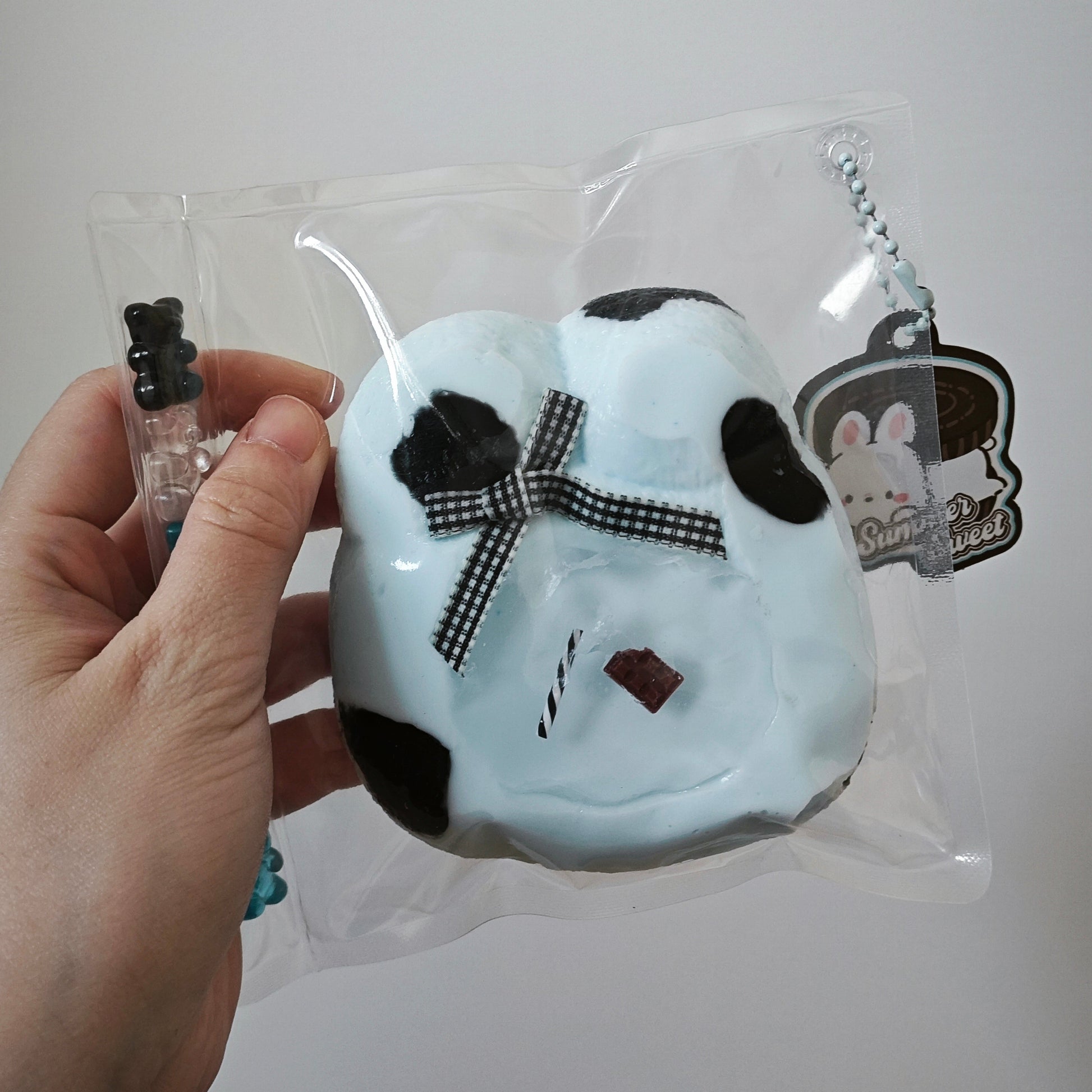 A hand holding a light blue squishy toy with black spots and a black-and-white checkered bow, sealed in a clear plastic bag with a keychain charm.