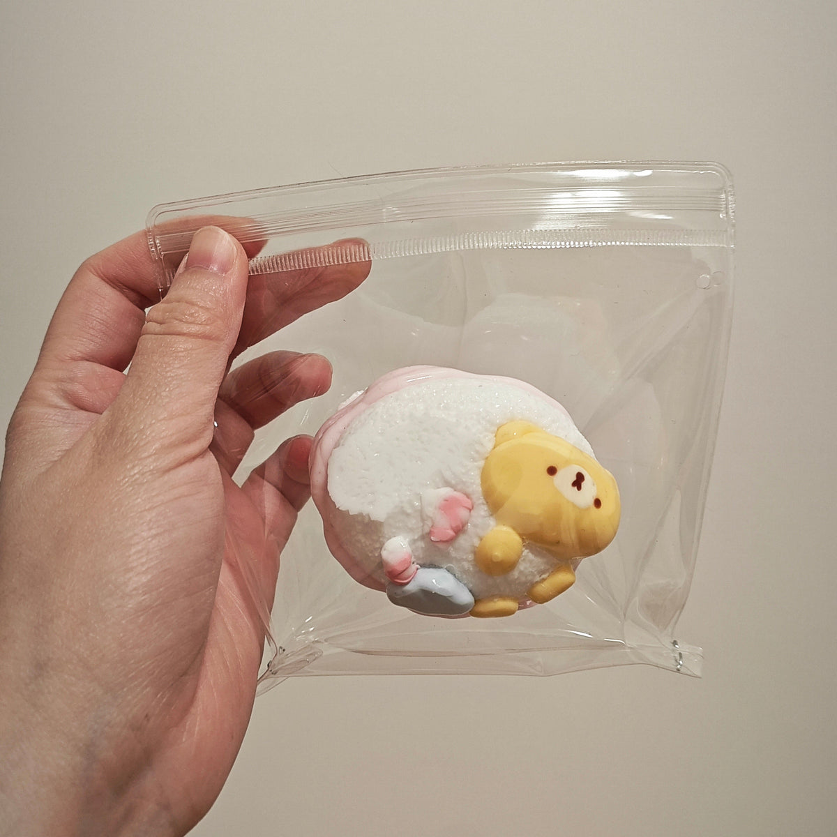 A hand holds a clear plastic zip-top bag containing a small plush toy with a white body, yellow face and legs, and tiny pink and blue accents, against a plain beige background.