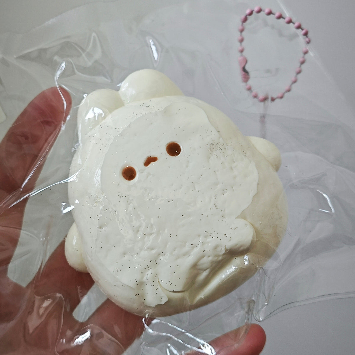 A hand holding a packaged white plush or foam character toy with a small pink beaded chain attached, shown against a plain light background.