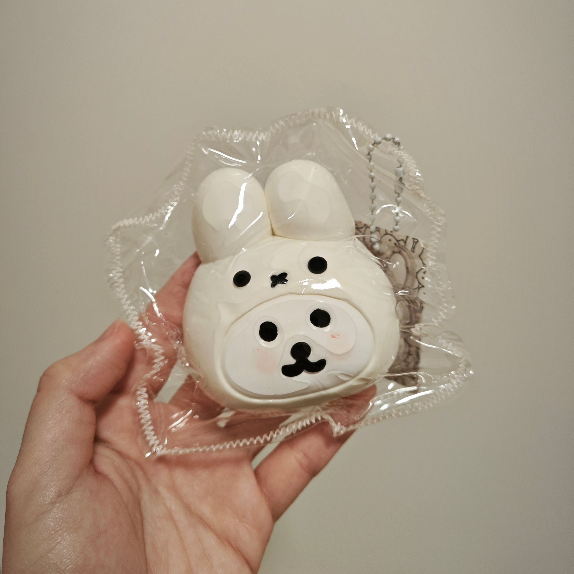 A hand holding a small white bunny-shaped accessory inside clear plastic packaging against a plain light beige wall.