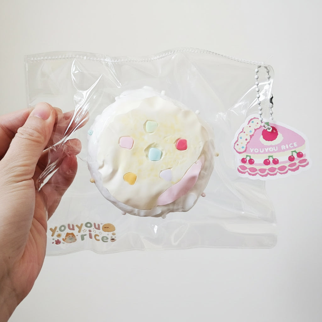 A hand holding a clear plastic bag containing a round, white, donut-like squishy toy with pastel-colored sprinkles. A small pink and white keychain charm attached to the bag reads 'YOU YOU RICE'.