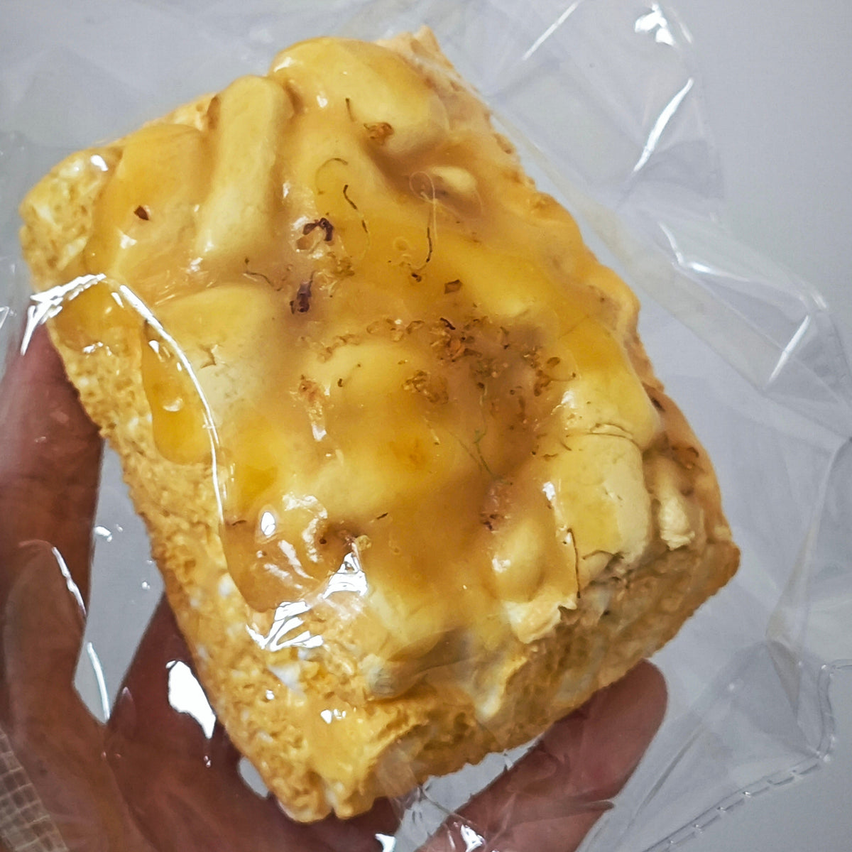 A square pastry or cake slice with a glossy golden-yellow glaze, held in a hand inside a clear plastic wrapper against a plain light background.