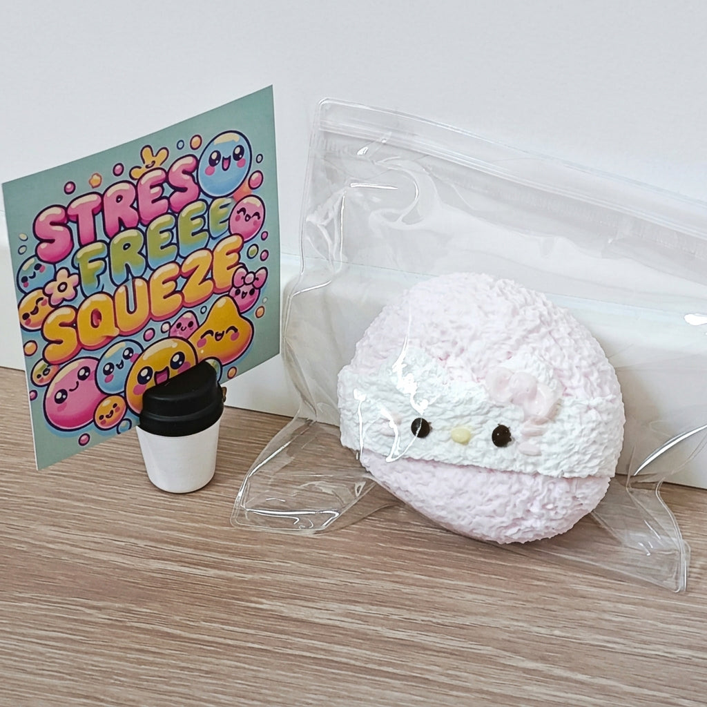 A pink plush squishy toy with a white band and small black eyes, sealed in a clear plastic bag next to a colorful 'STRESS FREE SQUEEZ' card and a small cup accessory on a wood surface against a white wall.