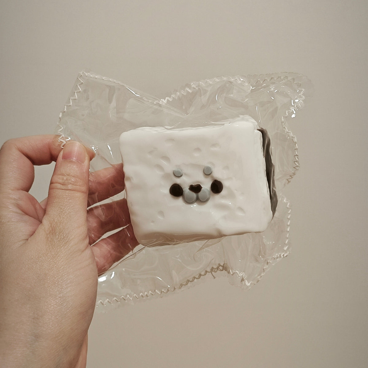 A hand holding a square white cake or pastry wrapped in clear plastic, decorated with a cute bear face made of dark brown and gray icing.