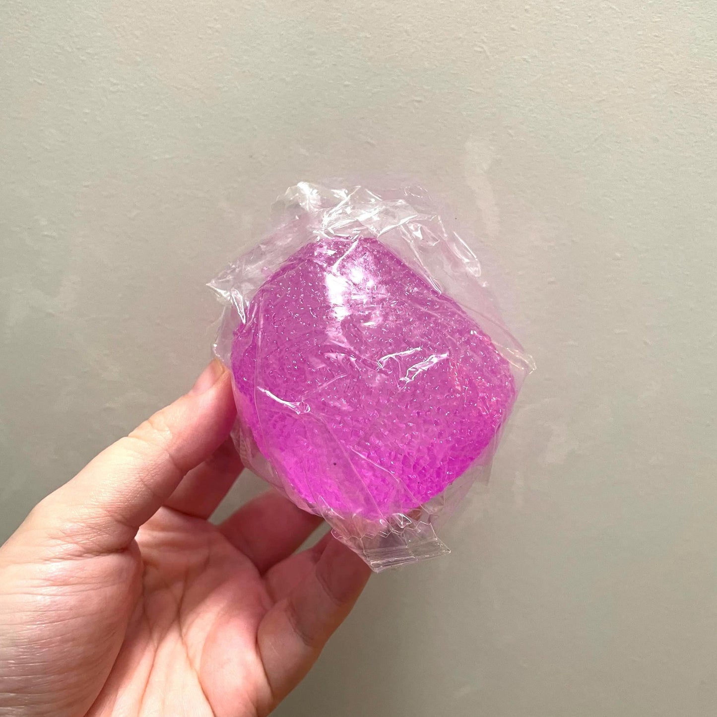 Hand holding a pink, translucent ball wrapped in clear plastic against a light gray background