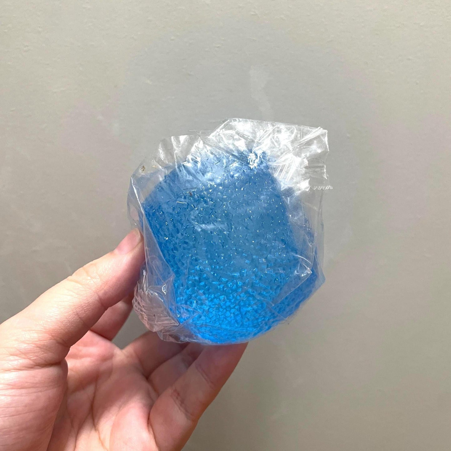 Hand holding a blue object wrapped in clear plastic against a neutral background