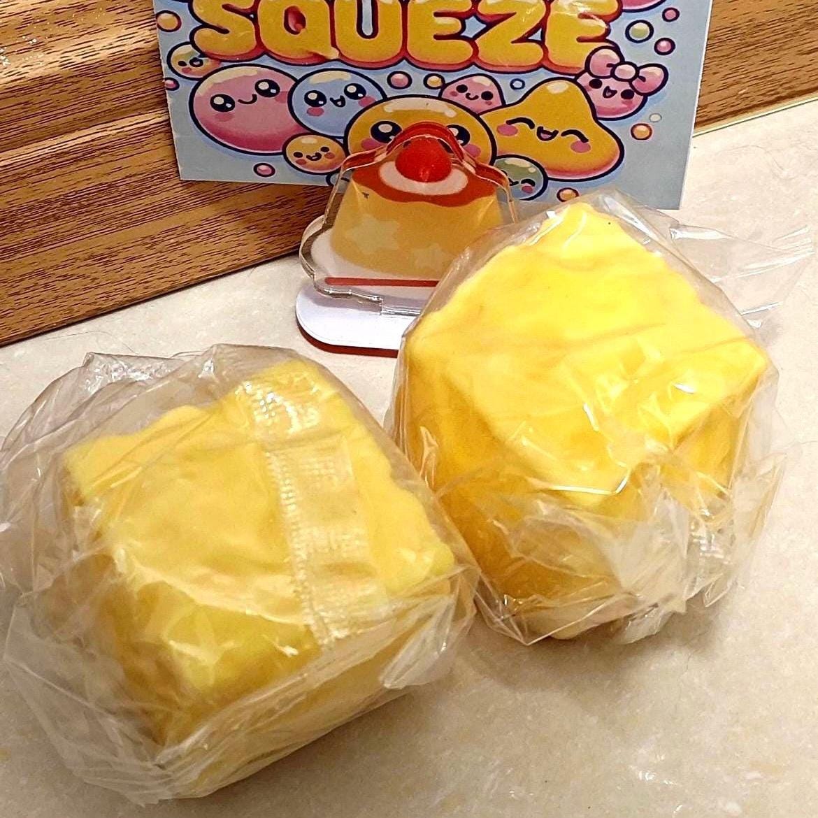 yellow, cube-shaped cheese wrapped in clear plastic. A card with the words "STRESS FREE SQUEEZE" and cartoon characters is in the background. A small, clear acrylic charm of a dessert is also visible.