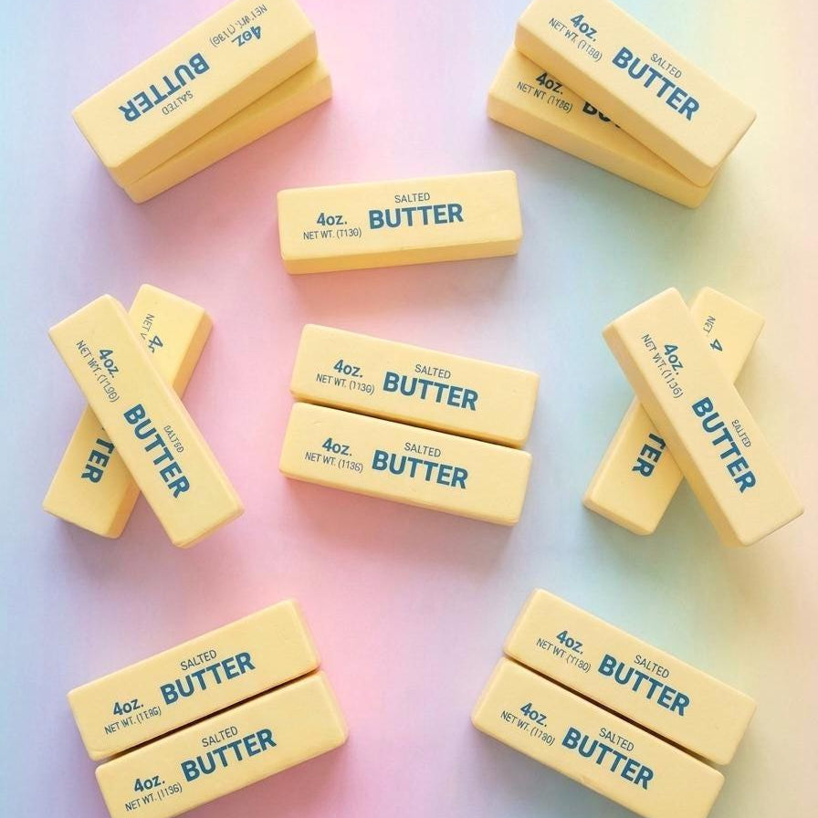 Twelve butter bars arranged in a flat-lay pattern on a pastel gradient background. Each bar is pale yellow with blue text reading '4 oz.