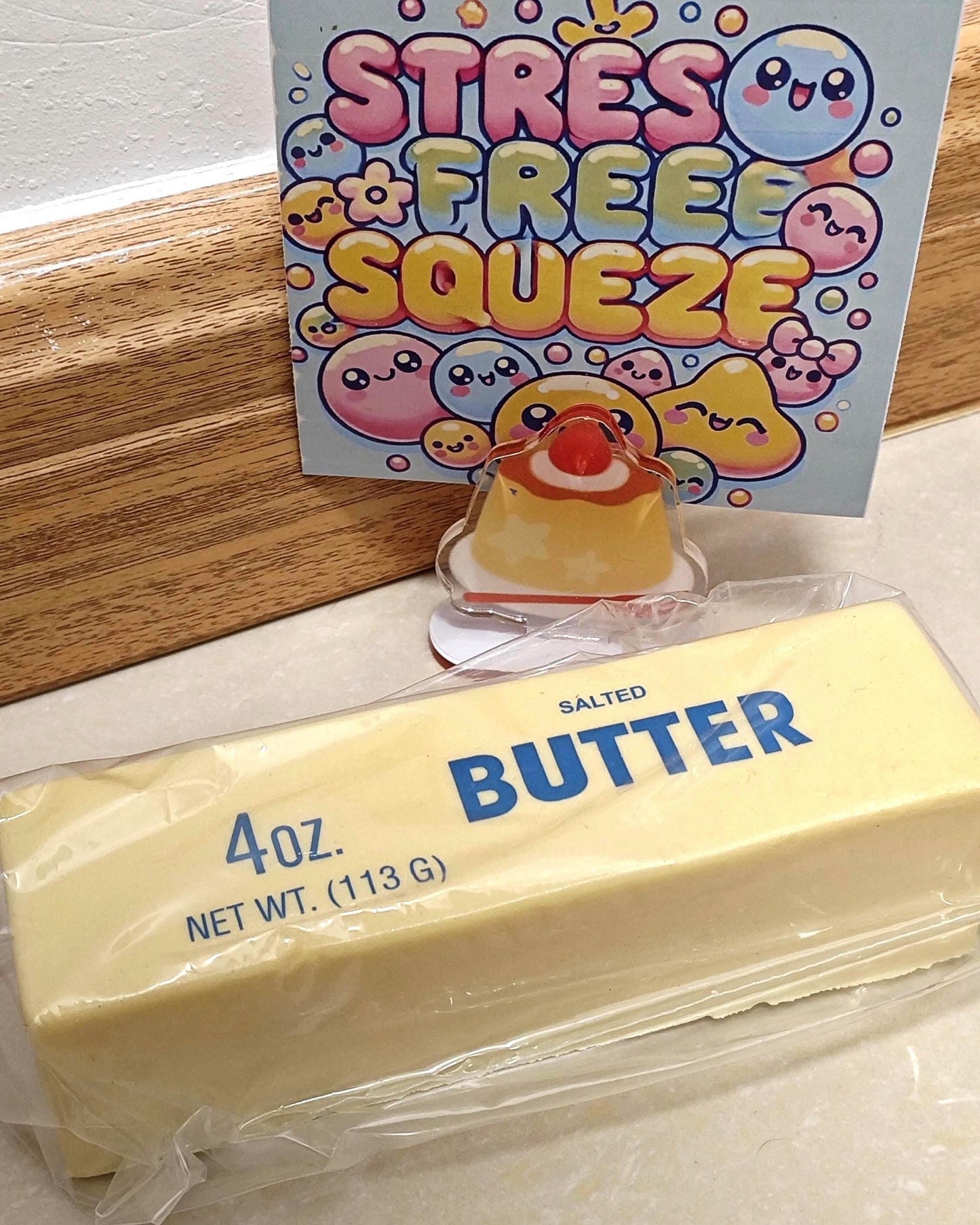 A stick of salted butter, in clear plastic packaging. A small, clear acrylic charm of a dessert with a red cherry on top. A card with the words "STRESS FREE SQUEEZE" in colorful bubble letters.