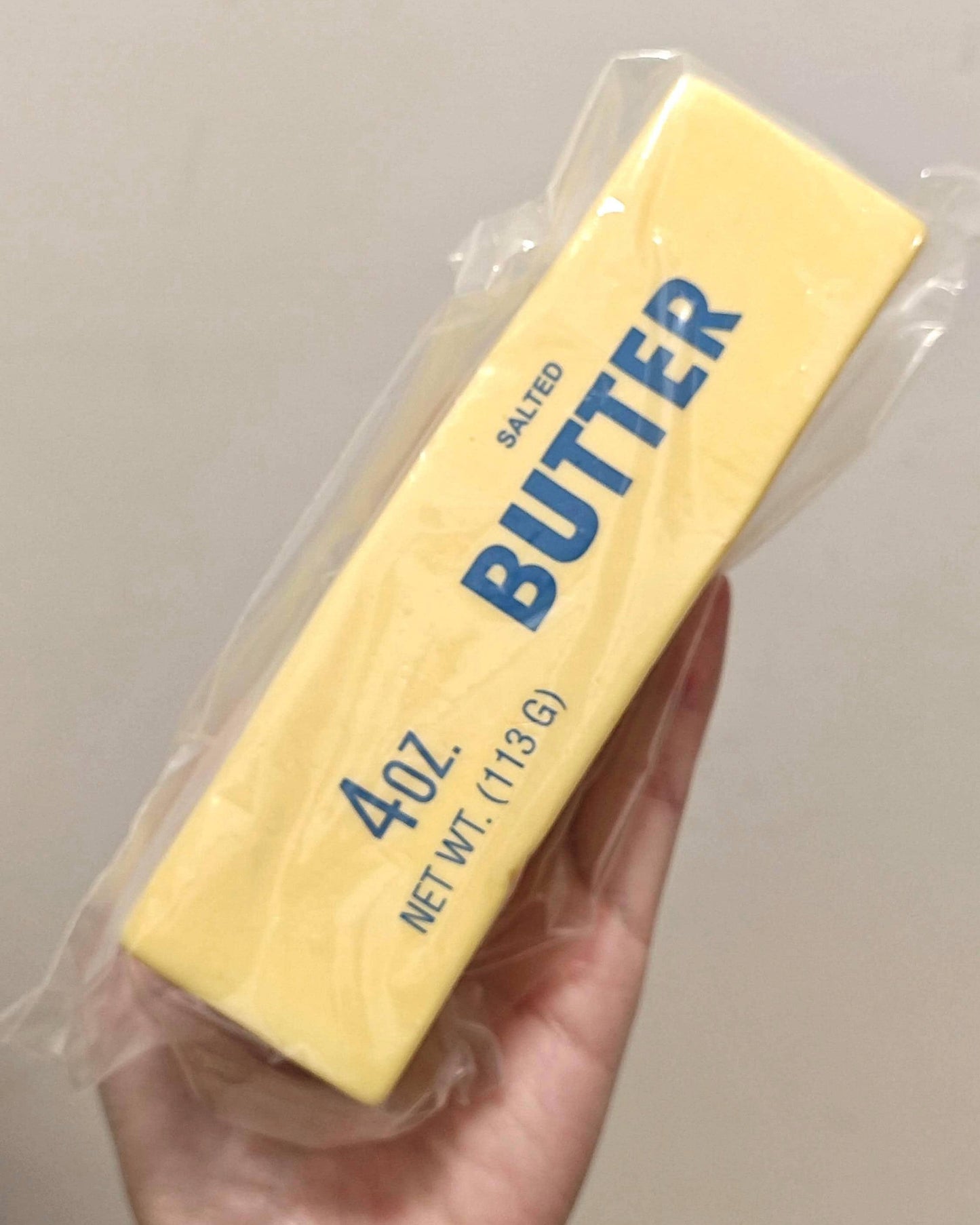 A hand holding a 4 oz stick of salted butter in its clear plastic wrapper, with 'BUTTER' printed in blue text on a pale yellow wrapper.