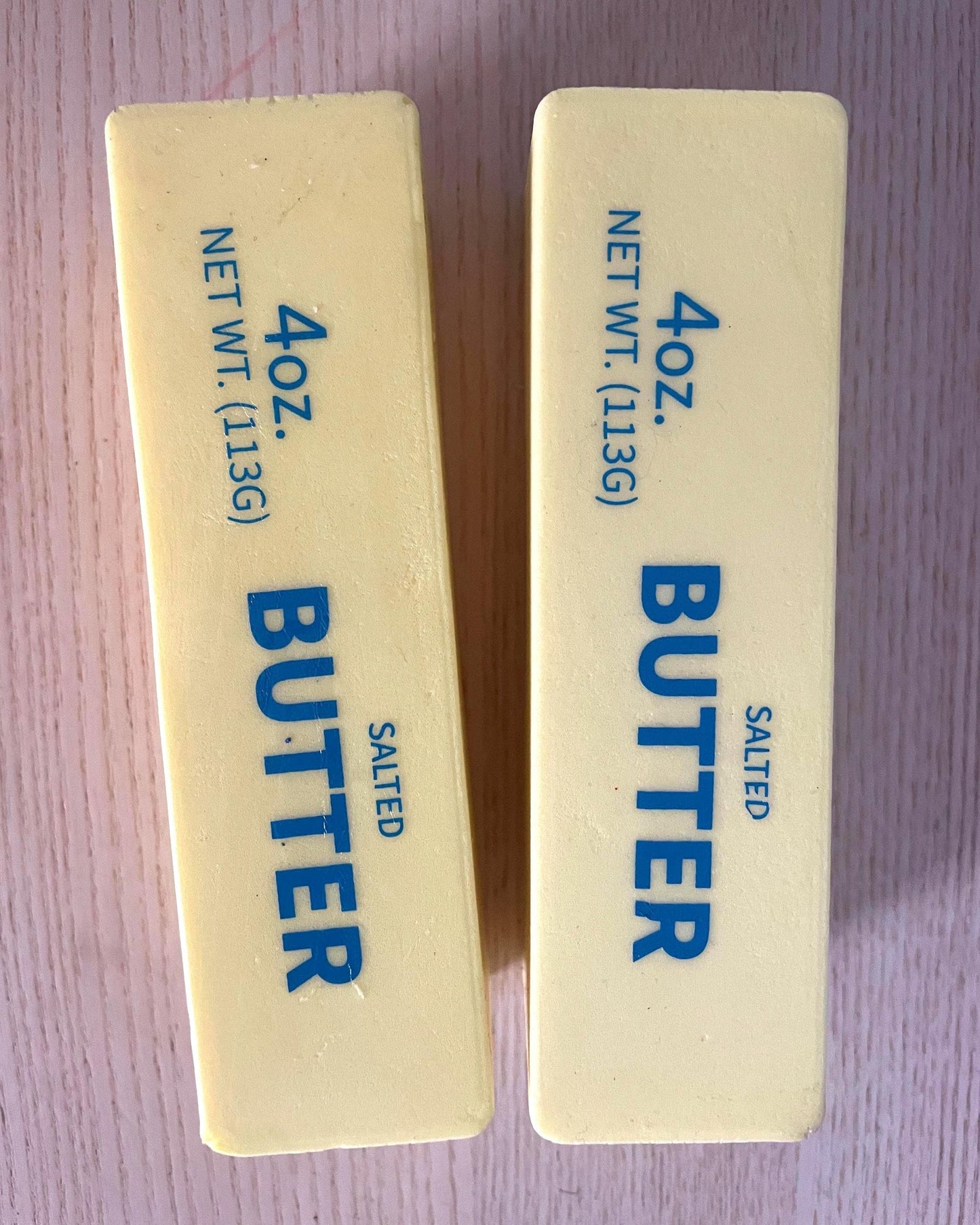 Two 4 oz sticks of salted butter standing upright on a light purple surface, with blue text printed on the pale yellow butter blocks.